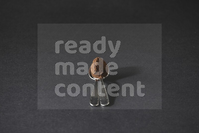 2 metal spoons on top of each other full of cloves powder on a black flooring