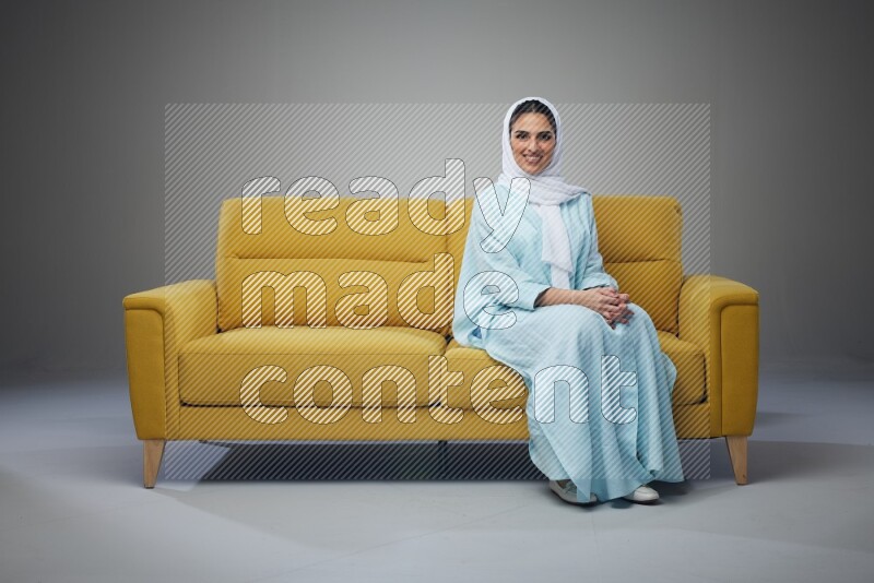 A Saudi woman wearing a light blue Abaya and white head scarf sitting on a yellow sofa and texting on her phone on a grey background