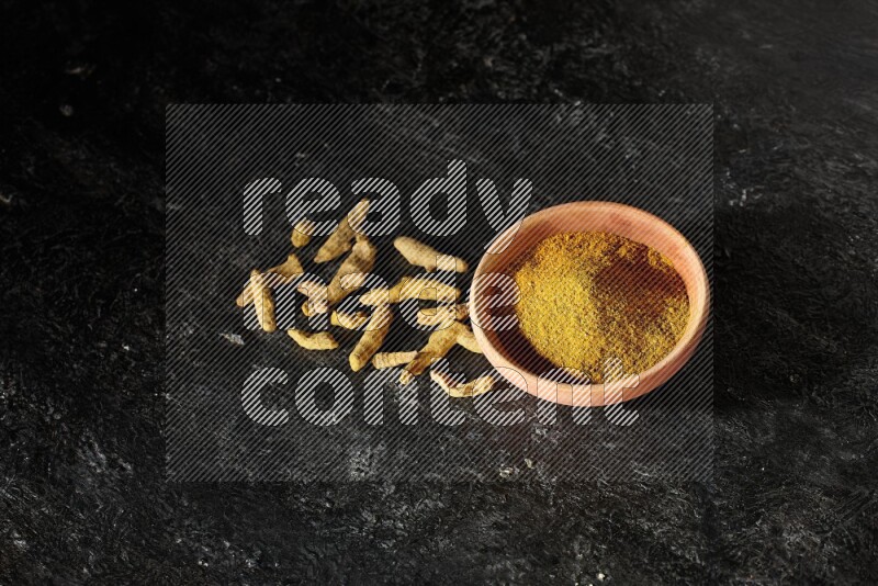 A wooden bowl full of turmeric powder with dried turmeric fingers on textured black flooring