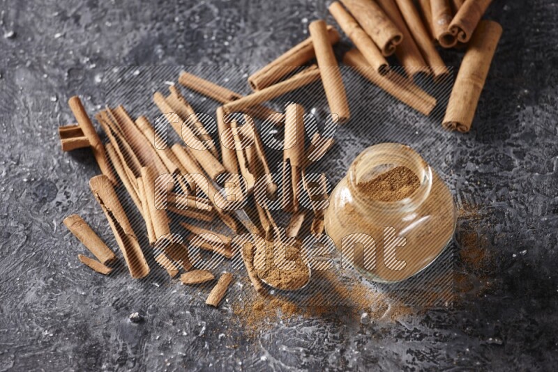 Herbal glass jar and a metal spoon full of cinnamon powder surrounded by cinnamon sticks on textured black background