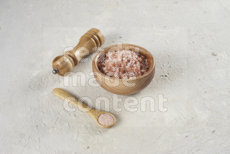 A wooden bowl and spoon filled with coarse pink himalayan salt and a wooden grinder beside them on white background
