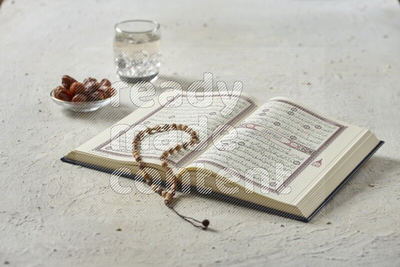 Quran with dates, prayer beads and different drinks all placed on textured white background