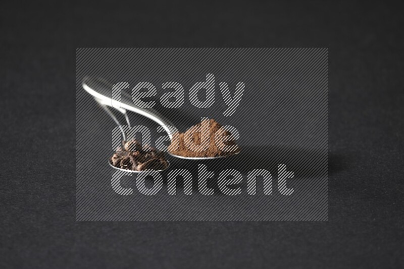 2 metal spoons, one full of cloves grains and the other full of cloves powder on a black flooring