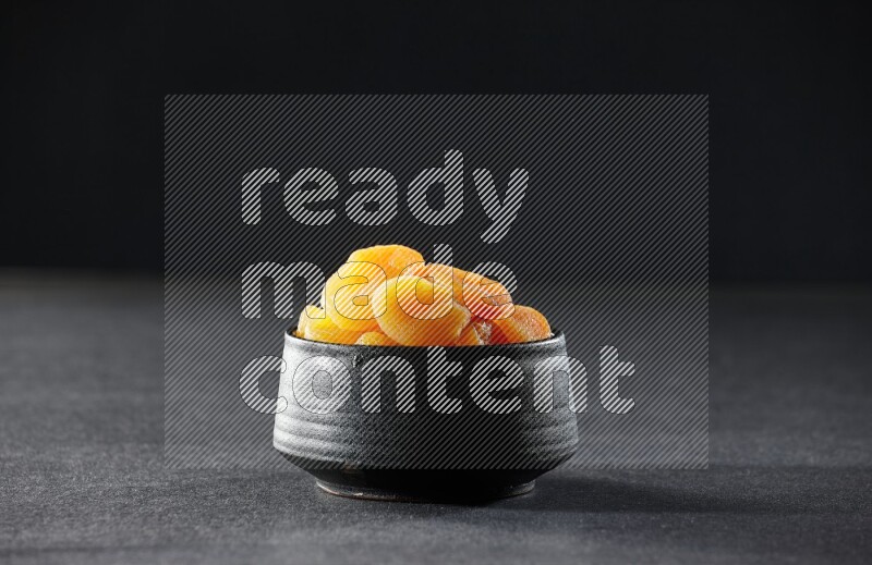 A black pottery bowl full of dried apricots on a black background in different angles
