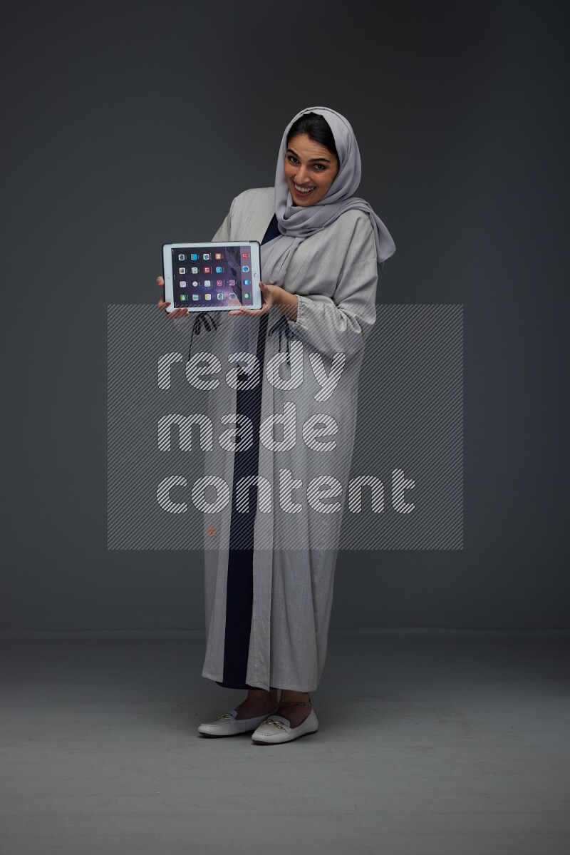 A Saudi woman wearing a light gray Abaya and head scarf standing and showing the phone's screen eye level on a grey background