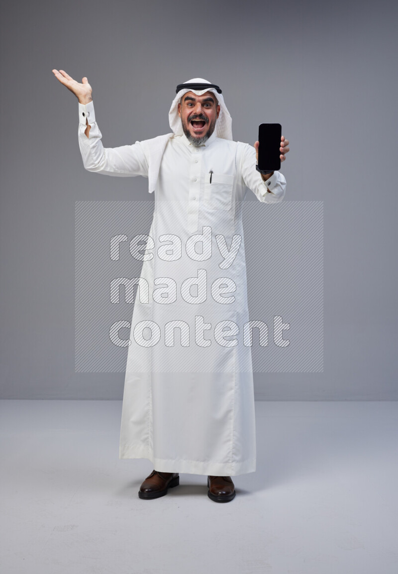 Saudi man Wearing Thob and white Shomag standing showing phone to camera on Gray background