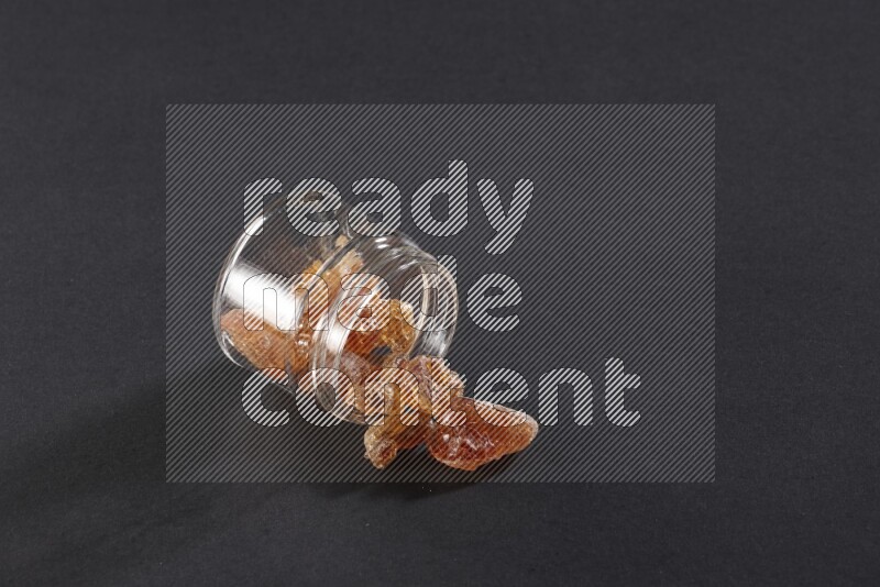 وعاء زجاجي ممتلئ بقطع الصمغ العربي والوعاء مقلوب وبعض القطع متناثرة خارجه علي خلفية سوداء