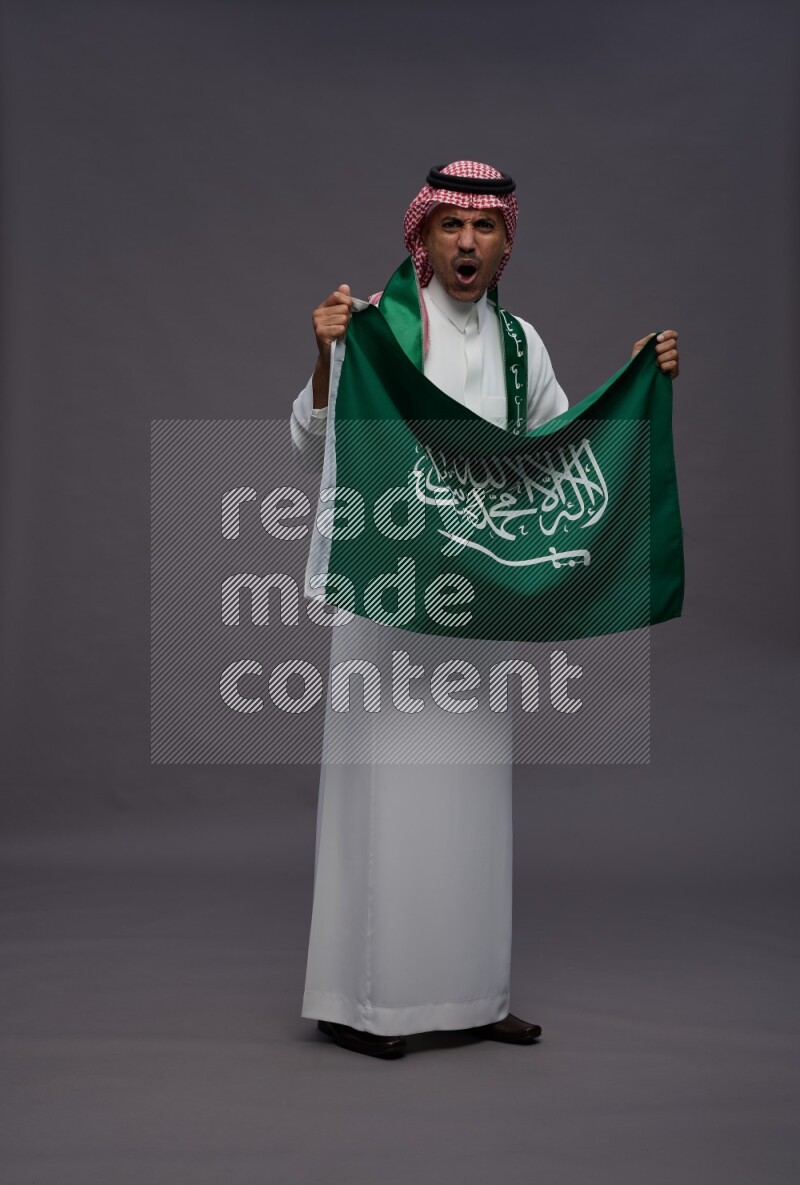 Saudi man wearing thob and shomag standing holding Saudi flag on gray background