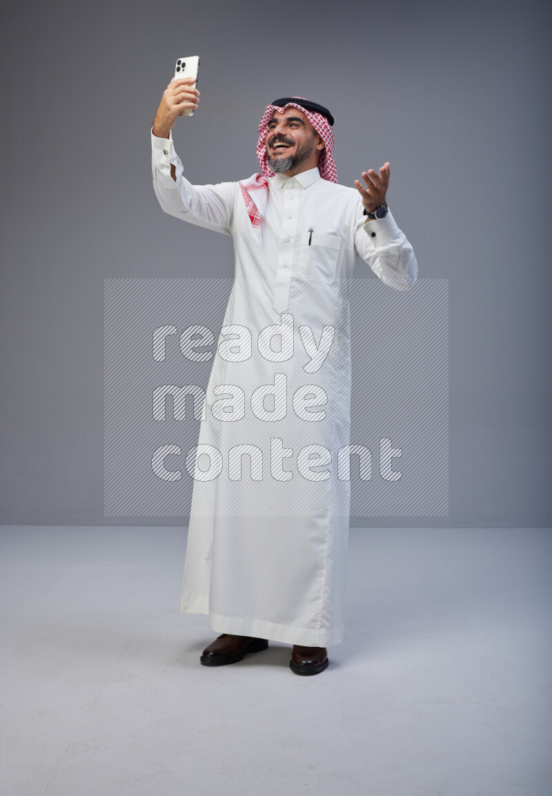 Saudi man Wearing Thob and red Shomag standing taking selfie on Gray background