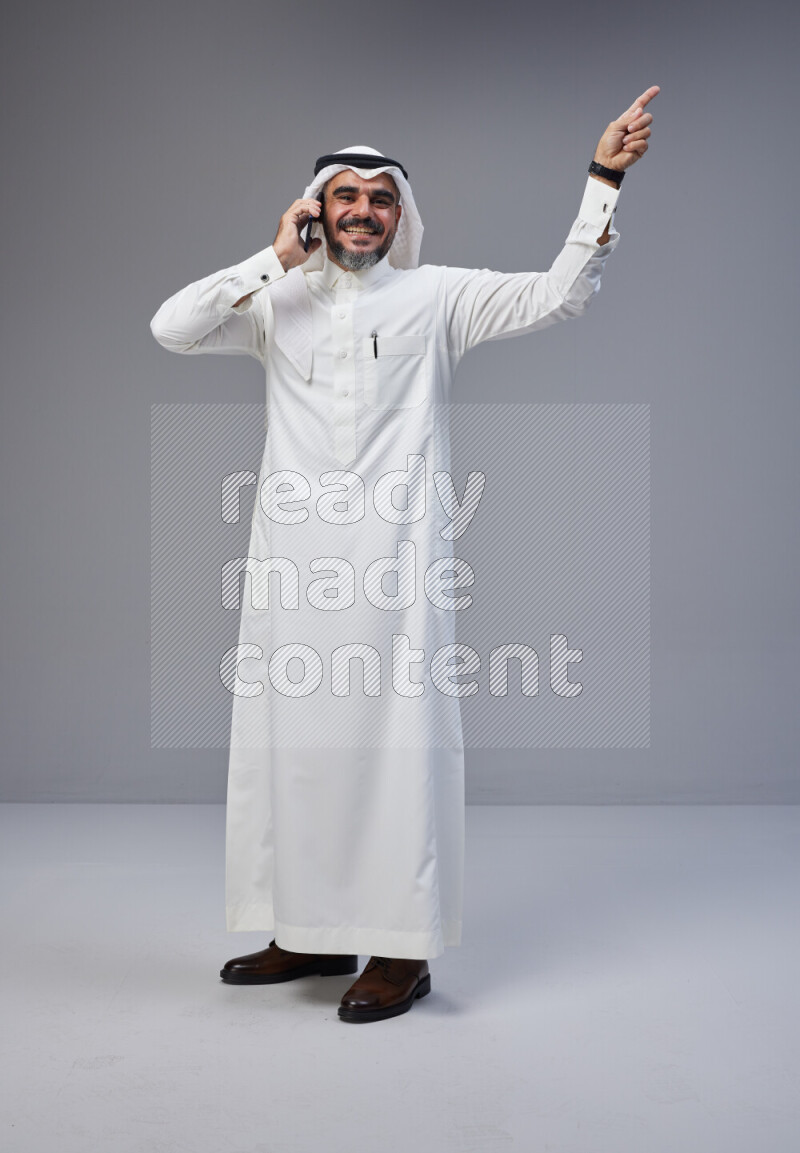 Saudi man Wearing Thob and white Shomag standing talking on phone on Gray background