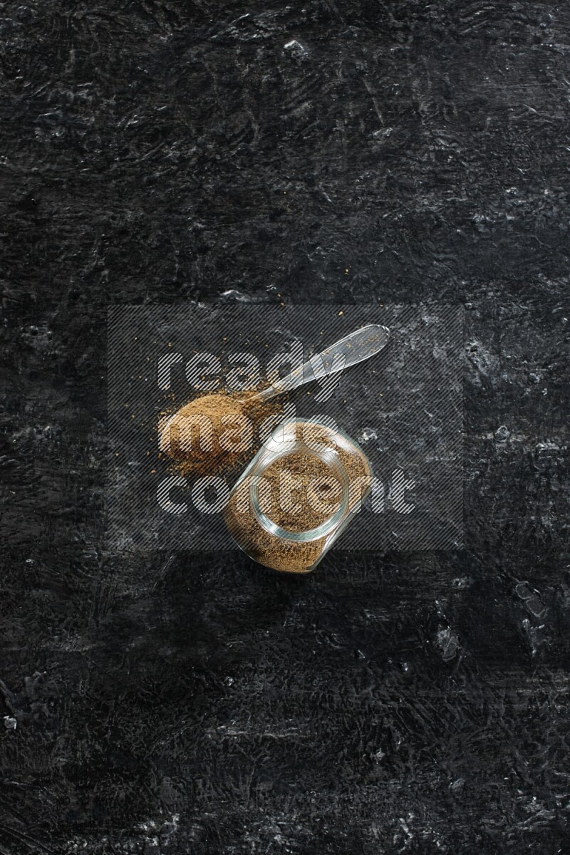 وعاء زجاجي للتوابل وملعقة معدنية ممتلئين ببودرة الكمون على خلفية سوداء