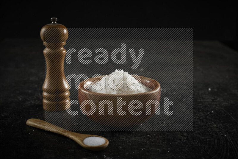 A wooden bowl and spoon filled with white sea salt and wooden grinder beside them on black background