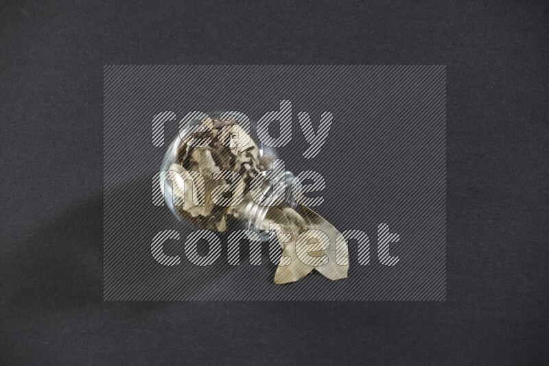 A glass spice jar full of dried bay leaves and jar is flipped and leaves spread out on black flooring