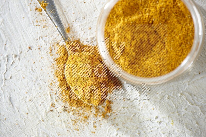 A glass jar and a metal spoon full of turmeric powder on a textured white flooring