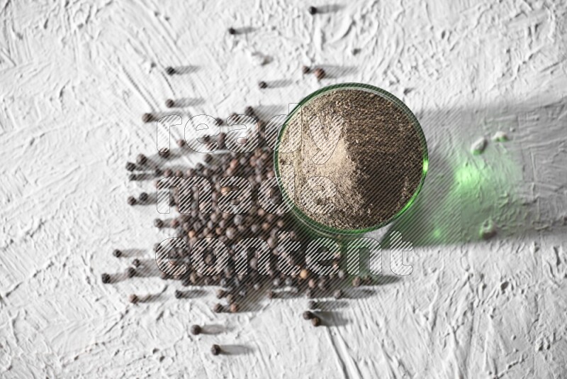 A glass cup full of black pepper powder and black pepper spread on a textured white flooring