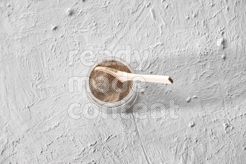 A glass jar full of black pepper powder with a wooden spoon on a textured white flooring