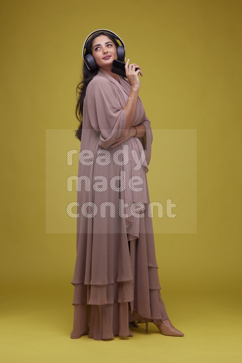 A Saudi woman Listing to Music on a Yellow Background wearing Brown Abaya