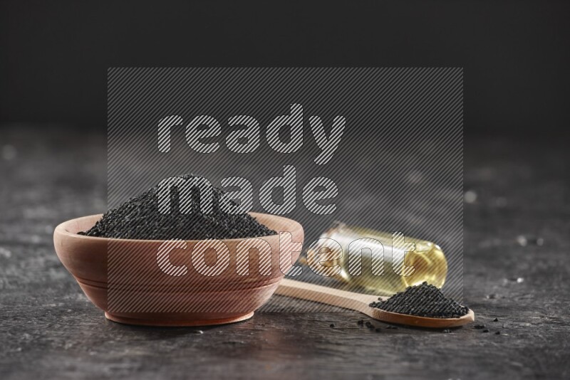 A wooden bowl and spoon full of black seeds and a bottle of black seeds oil on a textured black flooring