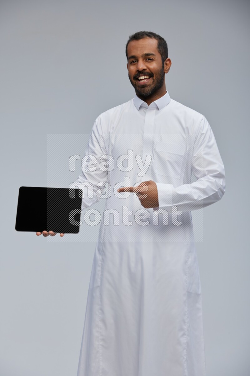 Saudi man wearing Thob standing showing tablet to camera on Gray background