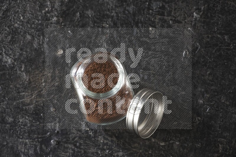 A glass spice jar full of cloves powder on textured black flooring