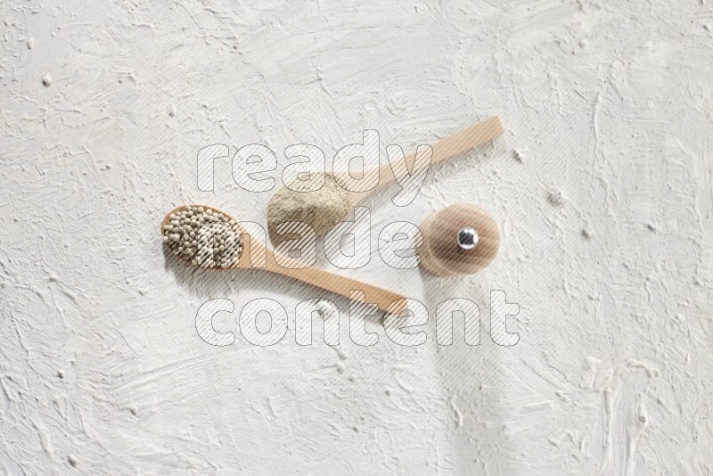 2 wooden spoons one full of white pepper powder and the other with pepper beads and a wooden pepper grinder on textured white flooring