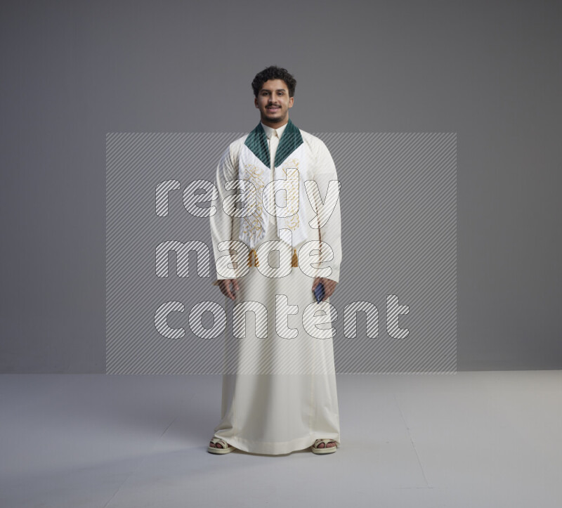 A Saudi man standing wearing thob and Saudi flag scarf on gray background