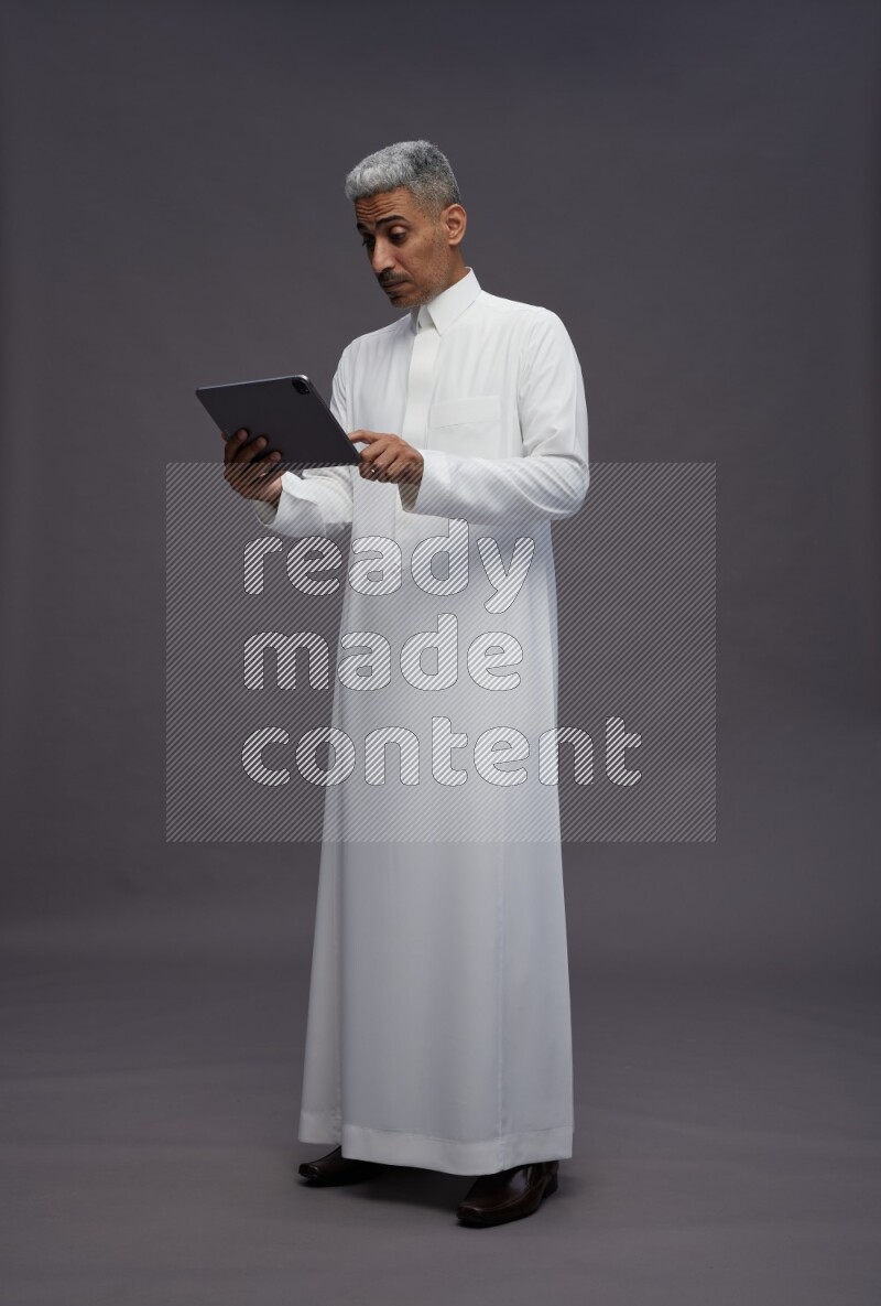 Saudi man wearing thob standing working on tablet on gray background