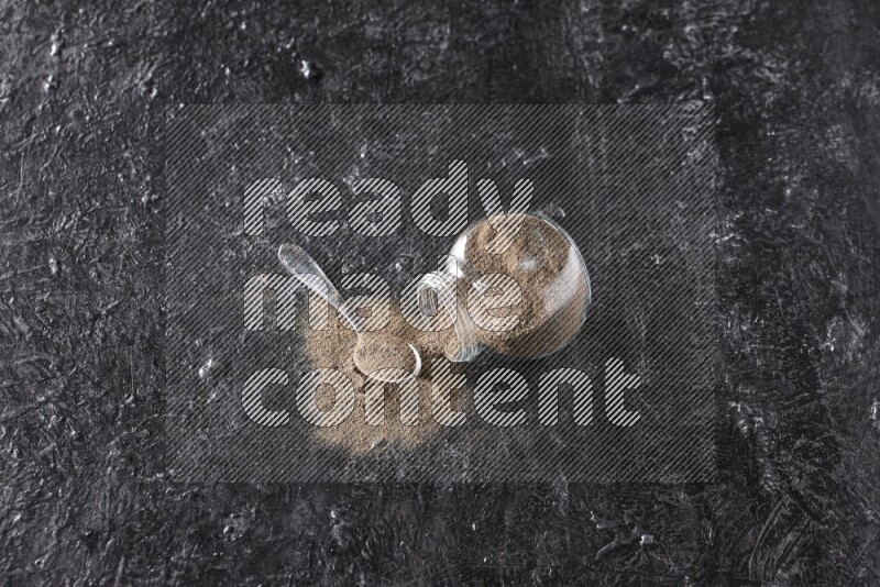 بودرة الفلفل الأسود في وعاء زجاجي مقلوب مع ملعقة معدنية مليئة بالبودرة علي خلفية سوداء