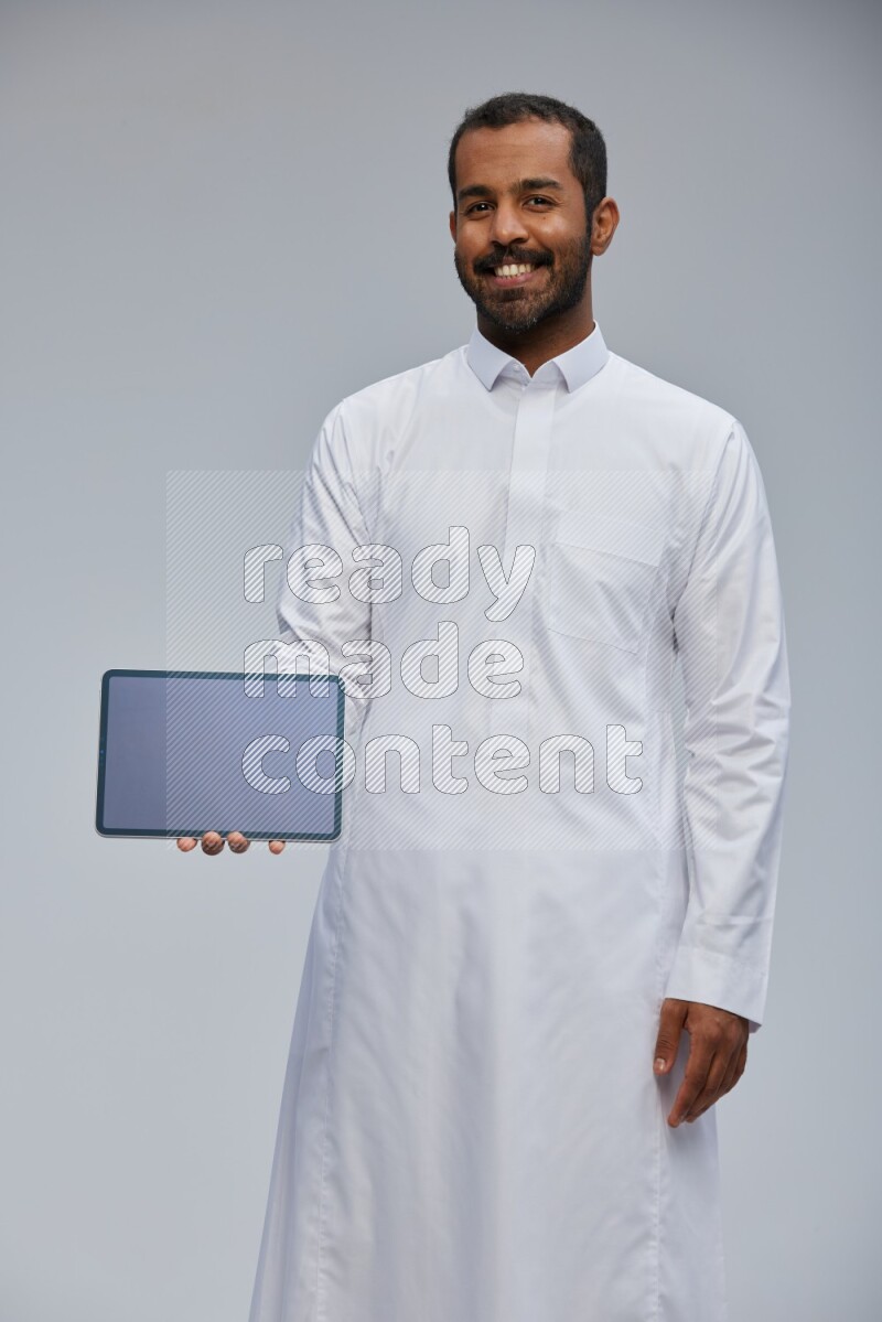 Saudi man wearing Thob standing showing tablet to camera on Gray background
