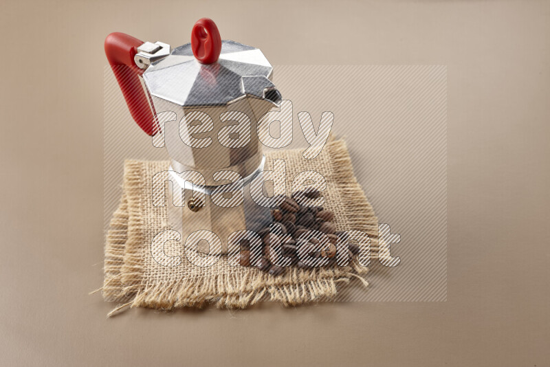 A moka pot with red handle surrounded by roasted coffee beans on beige background