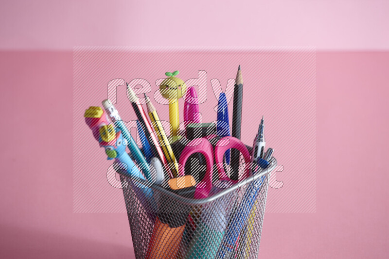 Pens, pencils and other different school supplies in a metal pen holder on rose background
