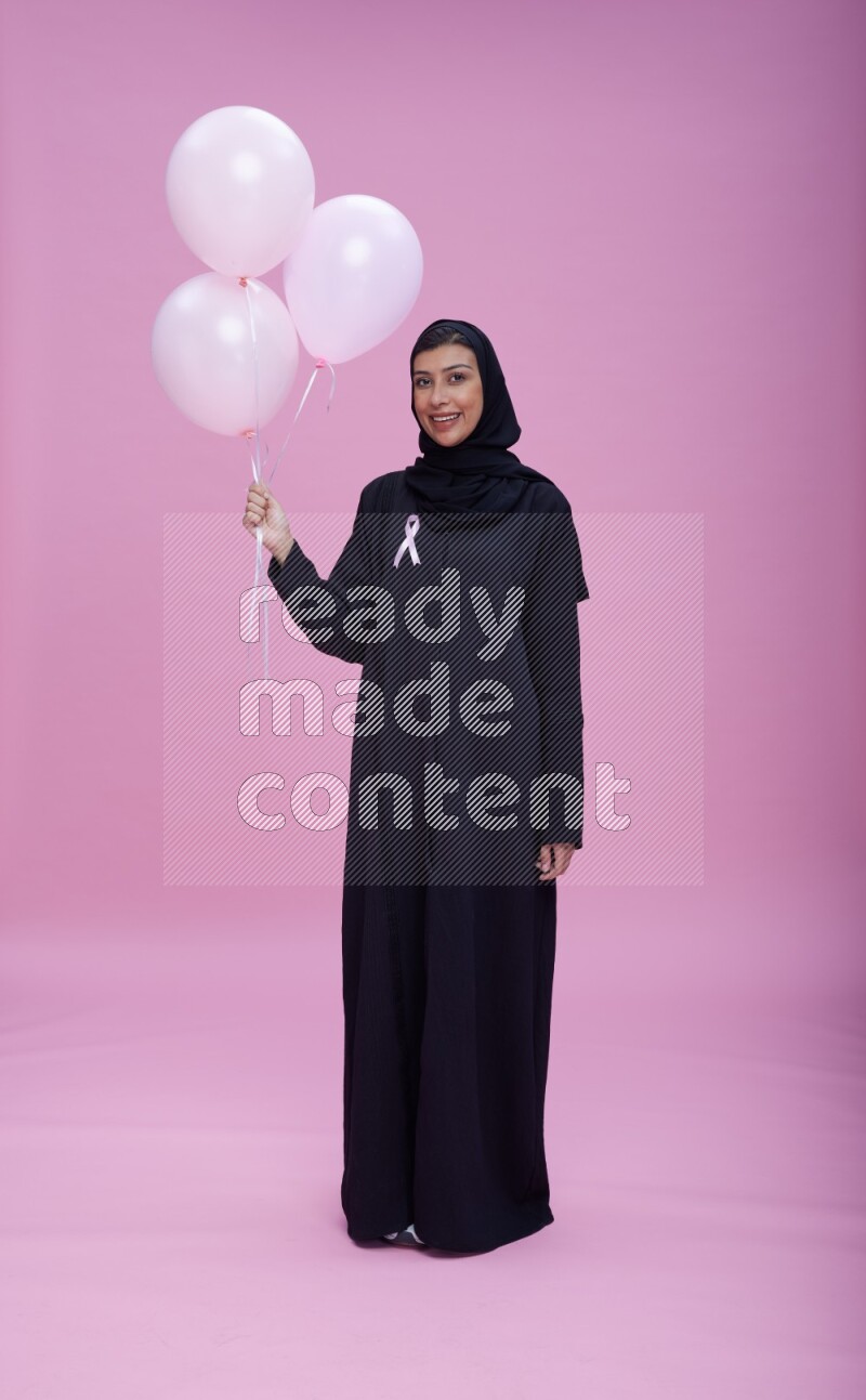 Saudi woman wearing pink ribbon on Abaya standing holding balloons on plain pink background