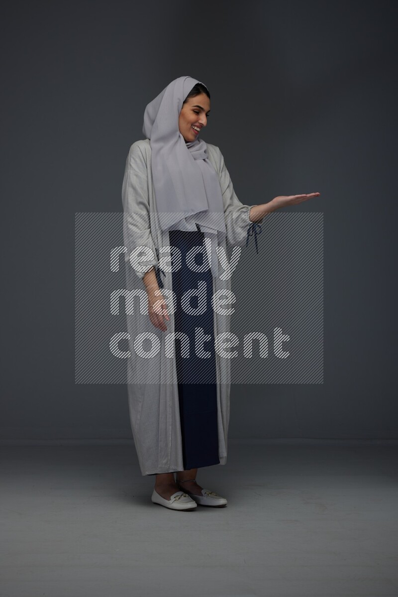 A Saudi woman wearing a light gray Abaya and head scarf standing and pointing in different directions eye level on a grey background