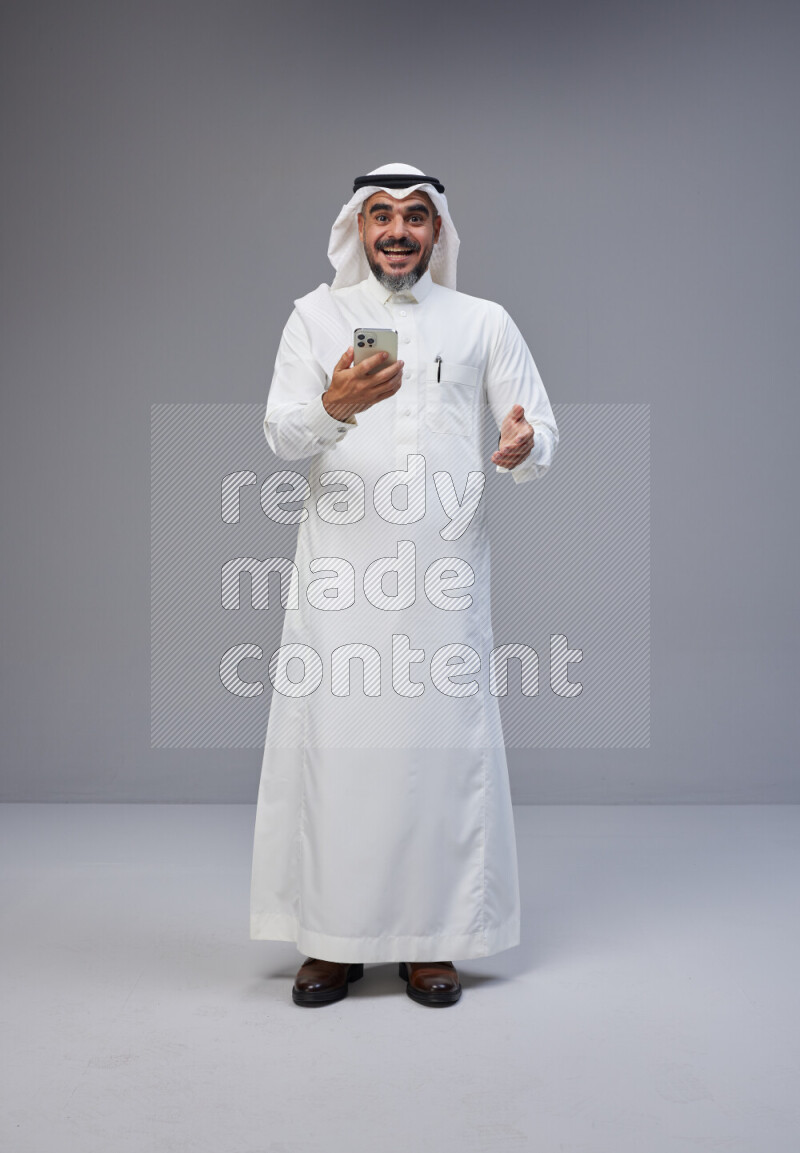 Saudi man Wearing Thob and white Shomag standing texting on phone on Gray background