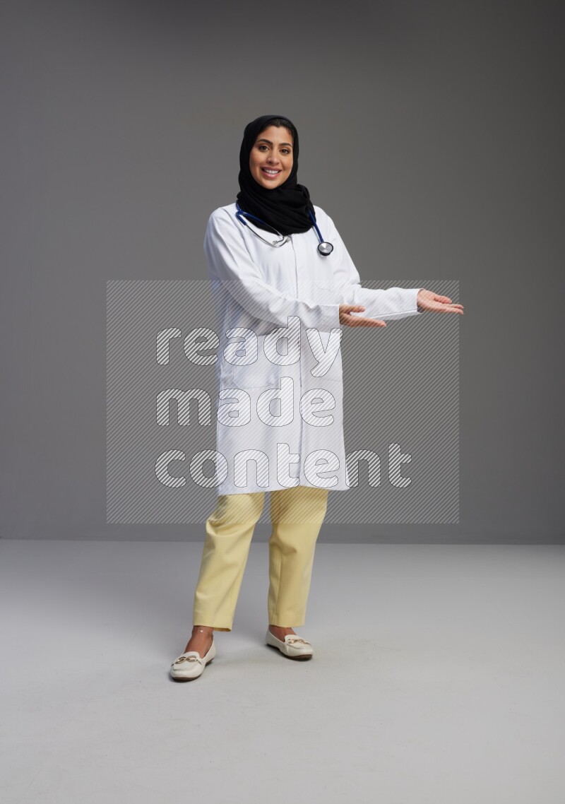 Saudi woman wearing lab coat with stethoscope standing interacting with the camera on Gray background
