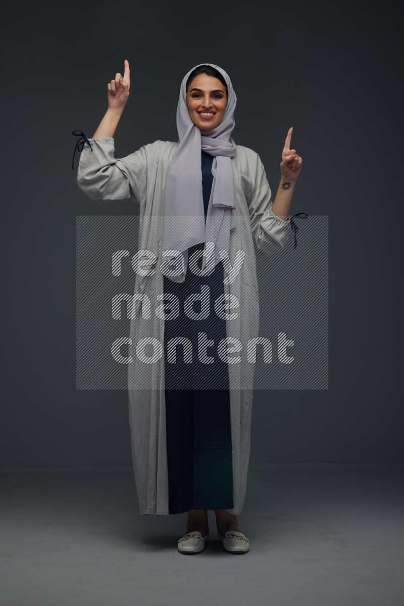 A Saudi woman wearing a light gray Abaya and head scarf standing and making multi poses eye level on a grey background