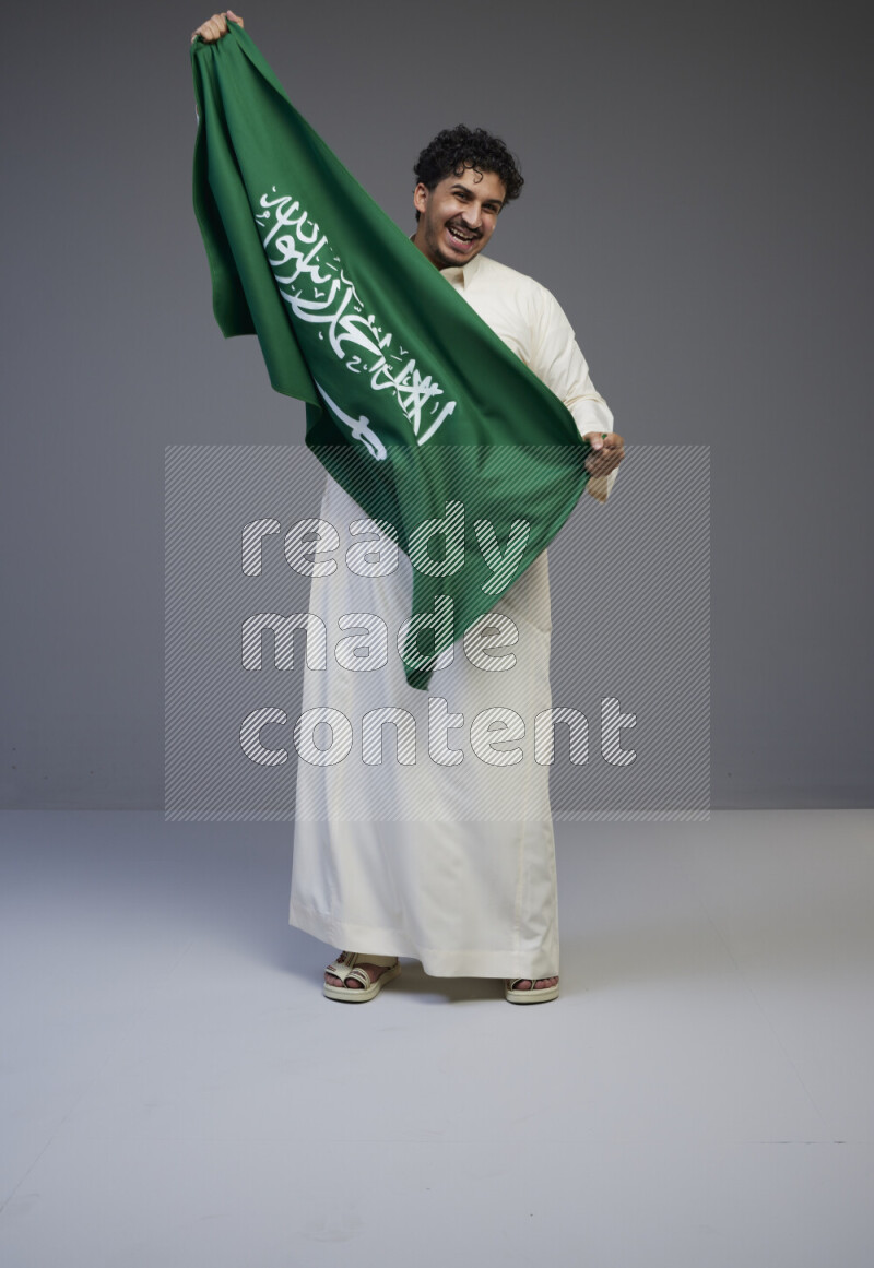 A Saudi man standing wearing thob holding big Saudi flag on gray background