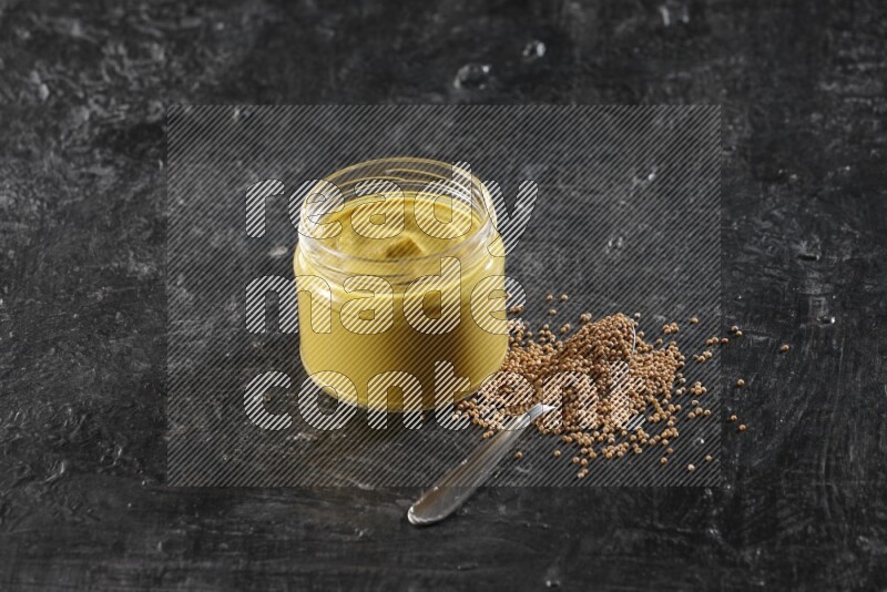 A glass jar full of mustard paste and a metal spoon full of mustard seeds on a textured black flooring