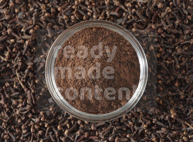 A glass bowl full of cloves powder and cloves grains spread on white flooring