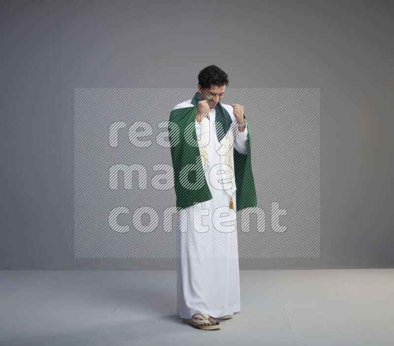 A Saudi man standing wearing thob with face painting wrapping Saudi flag scarf and holding big Saudi flag on gray background