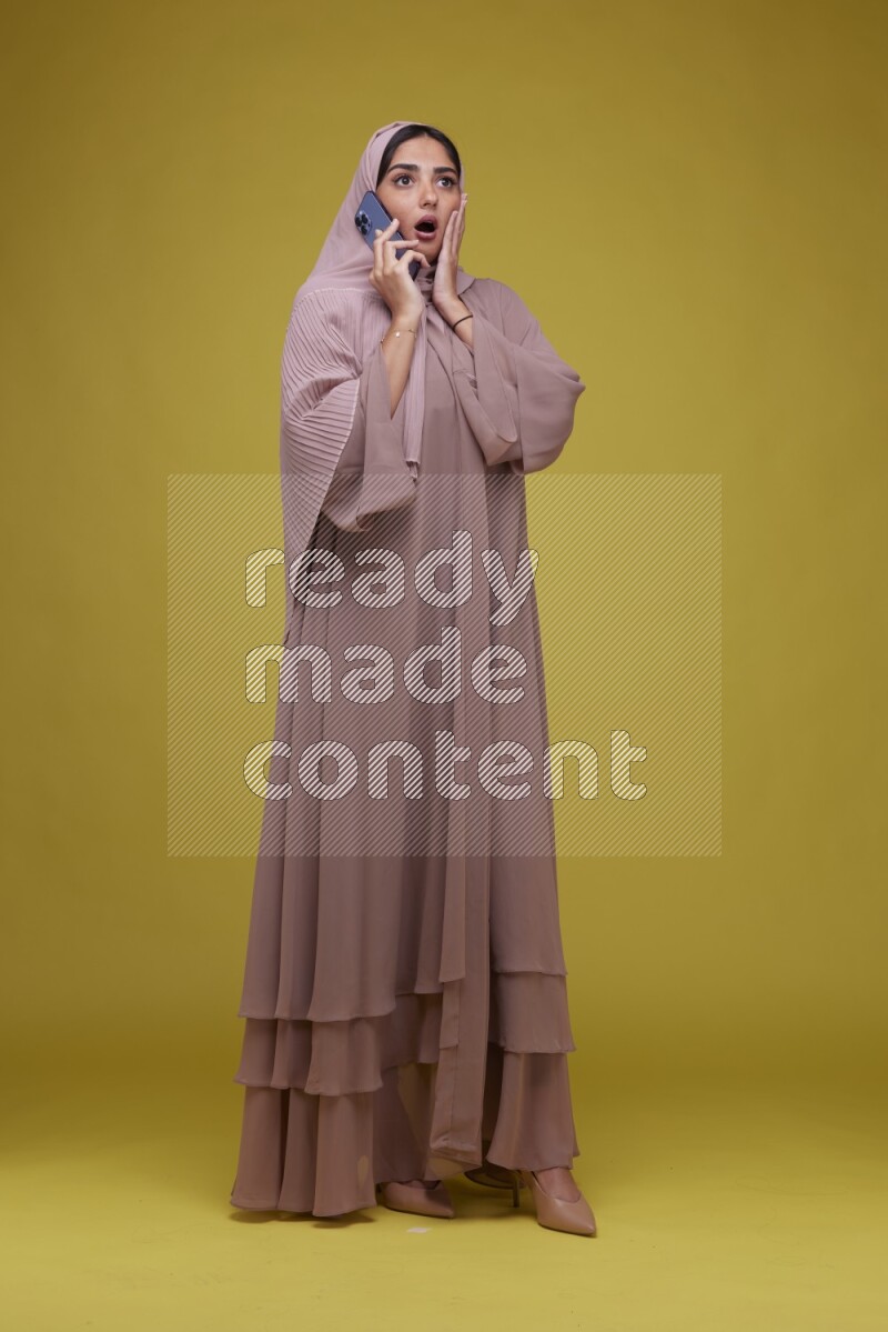 A woman having a call on a Yellow Background wearing Brown Abaya with Hijab