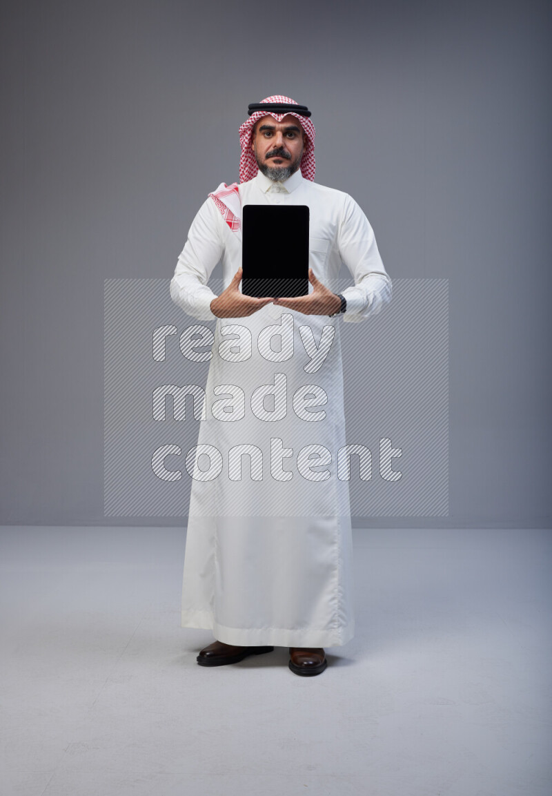 Saudi man Wearing Thob and red Shomag standing showing tablet to camera on Gray background