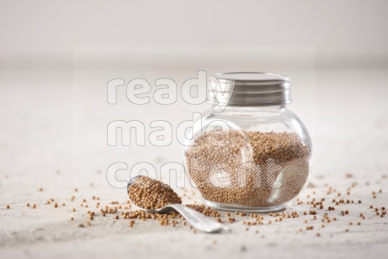 A glass spice jar and a metal spoon full of mustard seeds on a textured white flooring