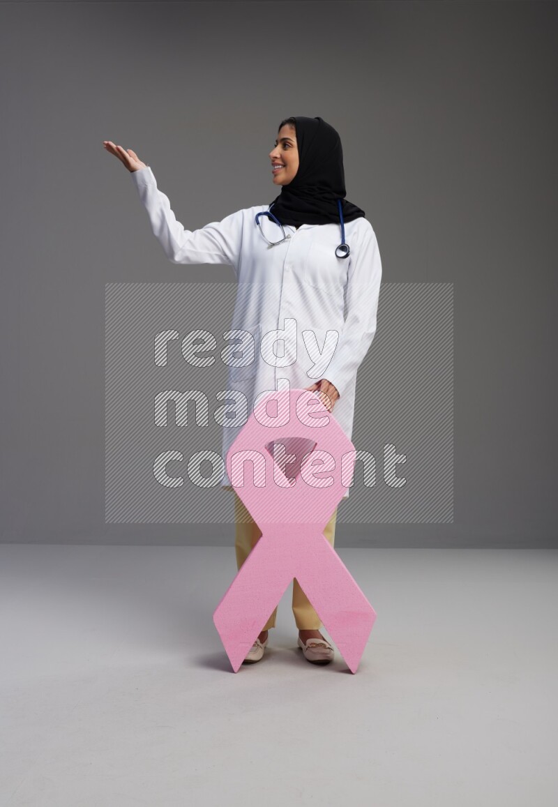 Saudi woman wearing lab coat with stethoscope standing holding awareness ribbon symbol standing on Gray background