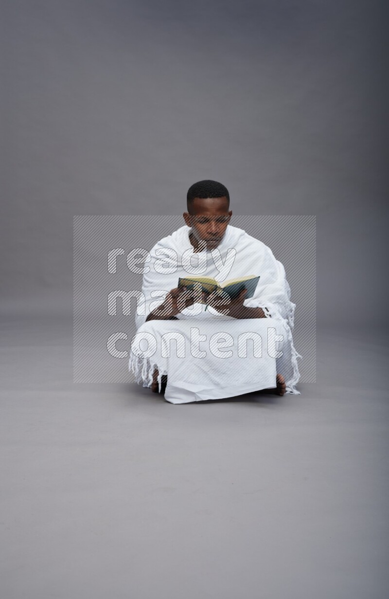 A man wearing Ehram sitting on floor reading quran on gray background