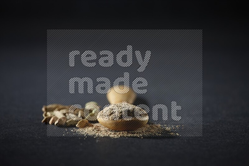 A wooden spoon full of cardamom powder and cardamom seeds beside it on black flooring