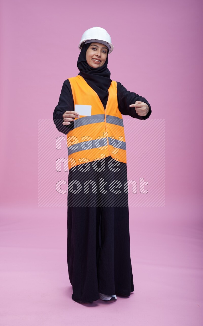 Saudi woman wearing Abaya with engineer vest and helmet standing holding ATM card on pink background