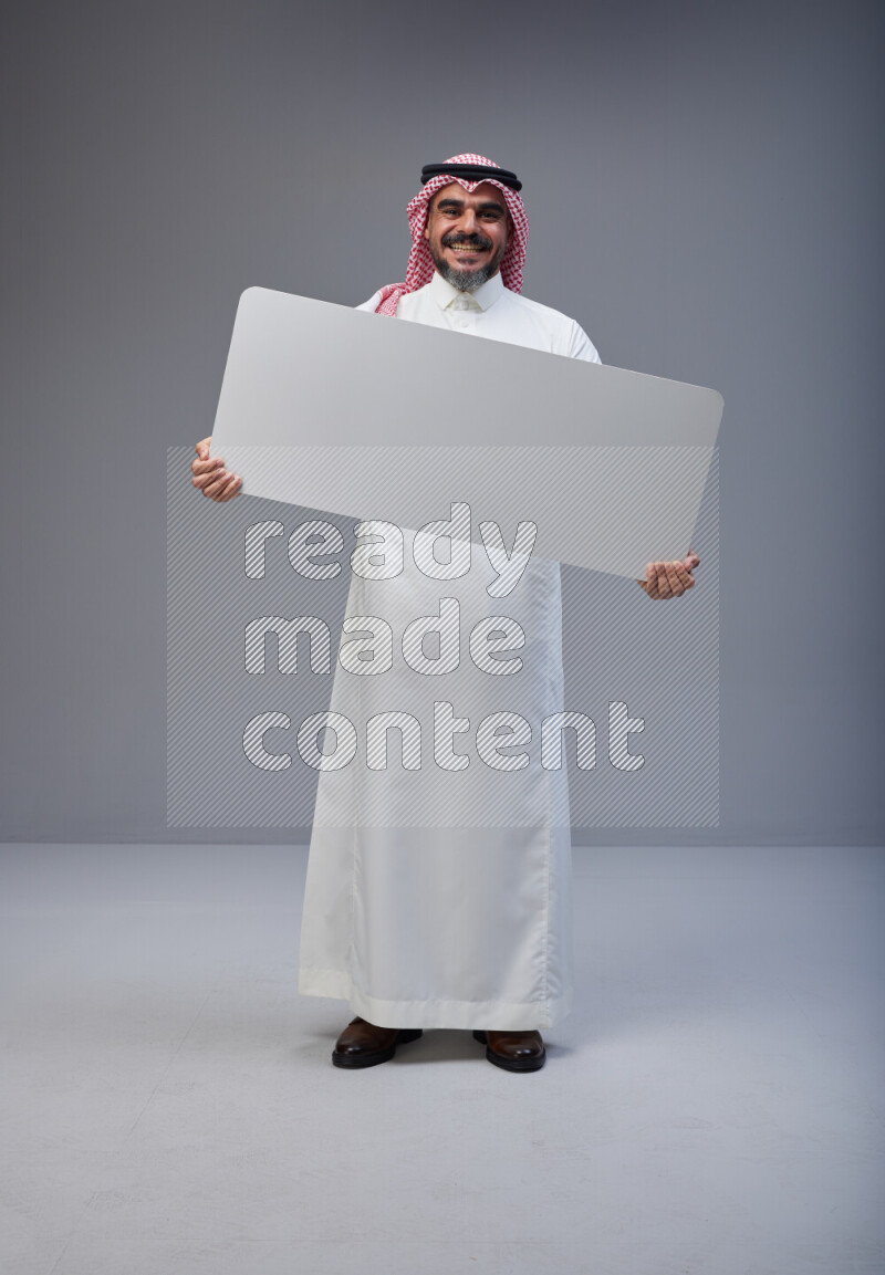 Saudi man Wearing Thob and red Shomag standing holding board on Gray background