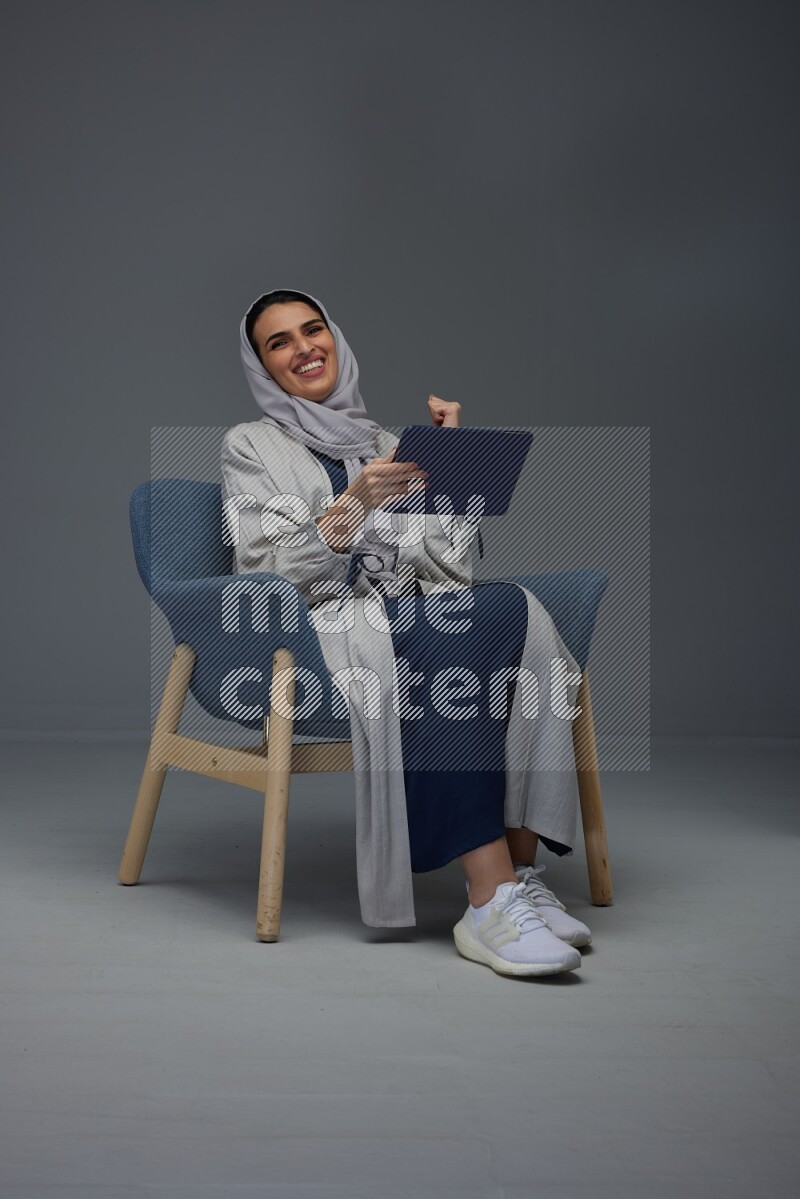 A Saudi female wearing a light gray Abaya and head scarf sitting on a dark grey chair while holding her iPad on a grey background