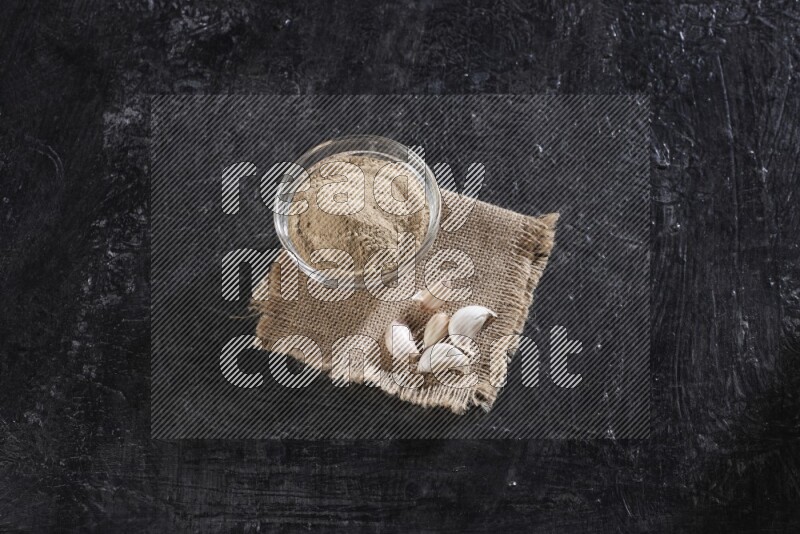 A glass bowl full of garlic powder placed on burlap fabric with garlic cloves on a textured black flooring